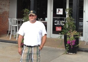 Man in plaid shorts stands outside a cafe.