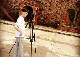 Man with camera tripod in front of building.