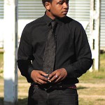 Man in a black suit and tie standing outside.