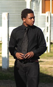 Man in a black suit and tie standing outside.