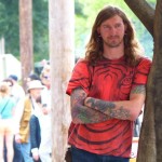 Man with tattoos and red shirt leaning against a tree.