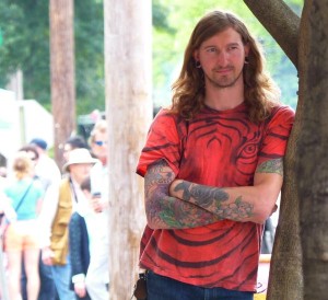 Man with tattoos and red shirt leaning against a tree.