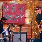 Man playing guitar with a drummer in front of a sign