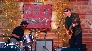 Man playing guitar with a drummer in front of a sign