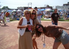 Three women smiling, holding drinks, outdoor festival.