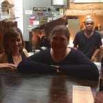 Three people at a bar with wine bottles.