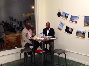 Two men talking at a table with photos on the wall.