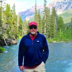 Man in blue shirt by a river in mountains.