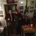 Four men in a room with a guitar on the wall.