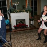 Man with microphone, woman in white dress by fireplace.