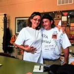 Two friends smiling, wearing white shirts with writing.