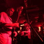 Man playing guitar on stage with red lighting.