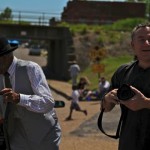 Two men, one holding a camera and the other a cane.