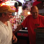 Two men at a bar, one wearing a white shirt.