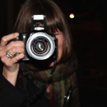 Woman holding a camera, looking into lens.