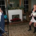 Man with microphone, woman in white dress by fireplace.