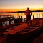 Man standing on dock with canoe at sunset.