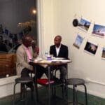 Two men talking at a table with photos on the wall.